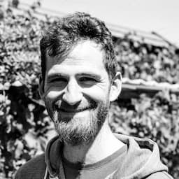 Black-and-white portrait of Sascha Zarhuber. It shows a man smiling towards the camera. He has a beard, and short, slightly curly hair. The background is blurred garden shed covered in some greenery.