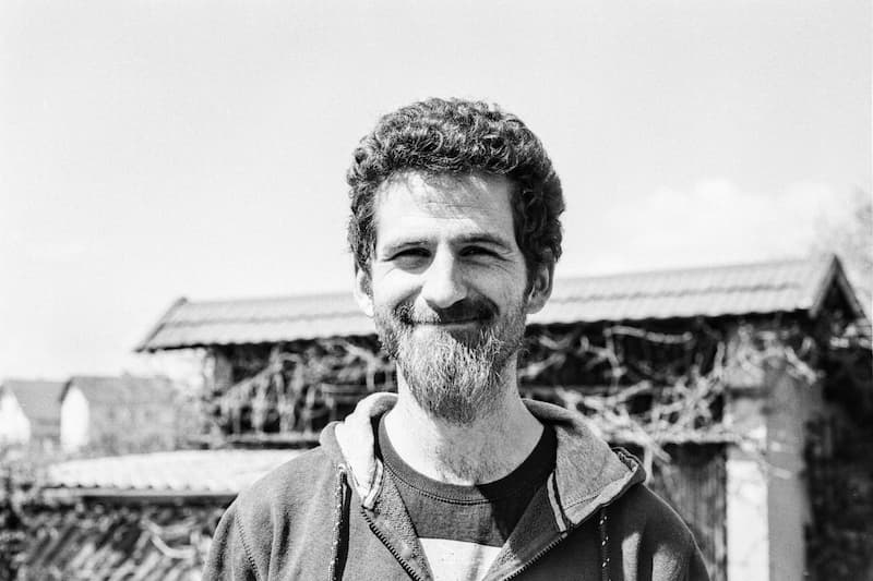 Black-and-white portrait of Sascha Zarhuber. He is wearing a hoodie and is standing in front of a blurry garden shed
            and smiling towards the camera.